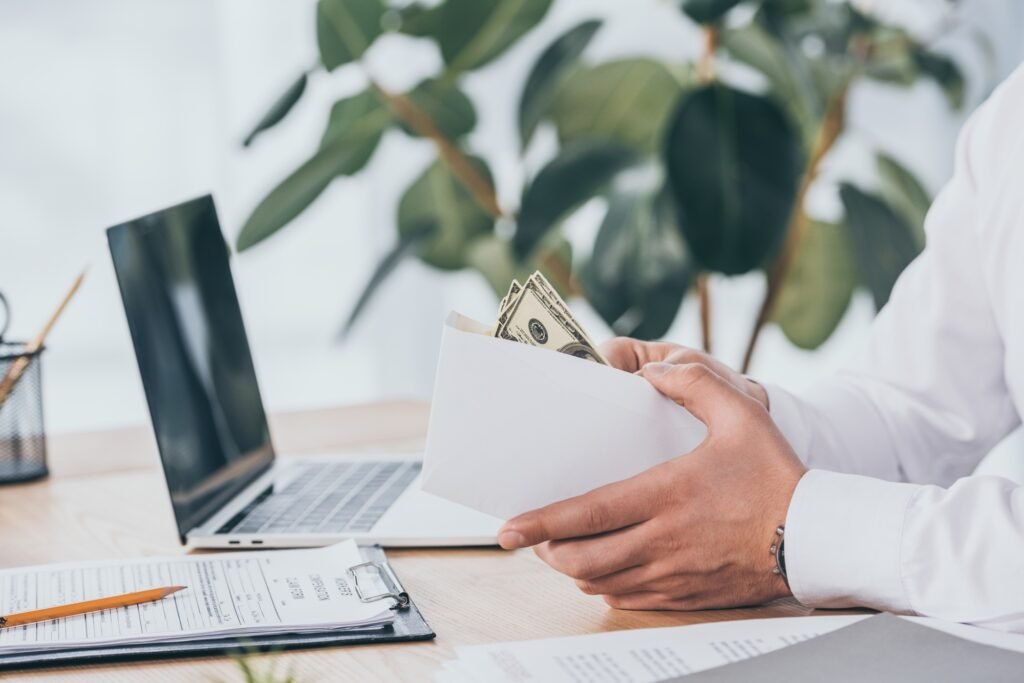 partial view of businessman holding envelope with money in hands at workplace, compensation concept